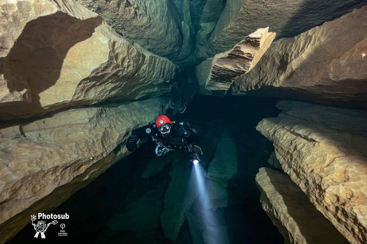 Cave diver with red helmet and primary light descending through a narrow vertical crack between rock walls