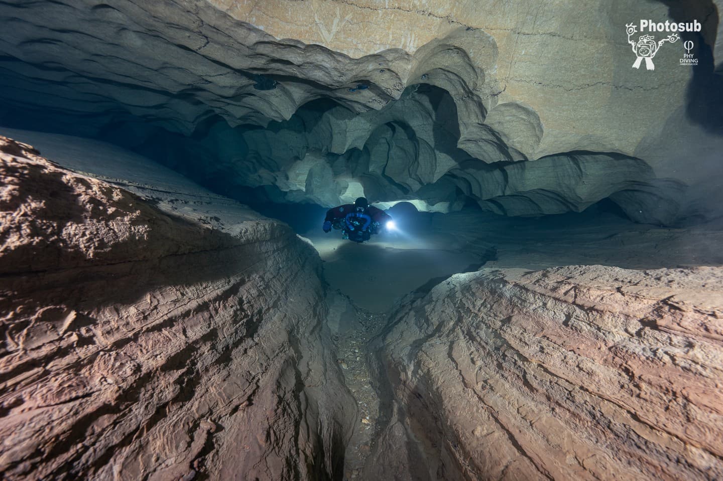 Diver in a low cave passage between layered rock formations with sandy floor