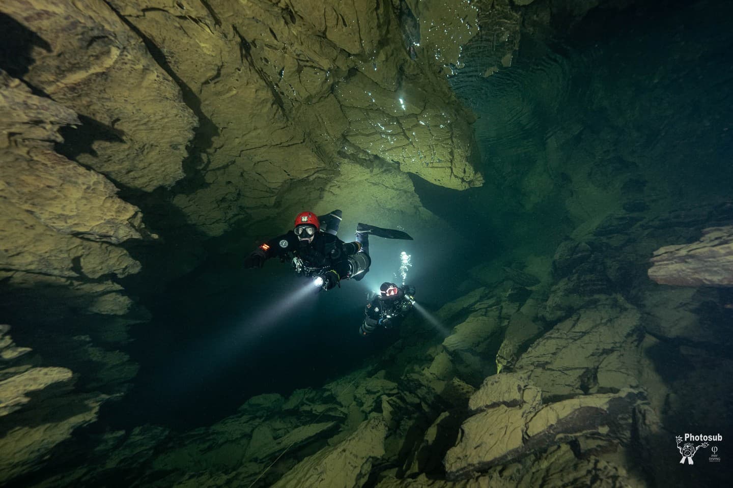 Two cave divers swimming through rocky cave passage with primary lights illuminating the way