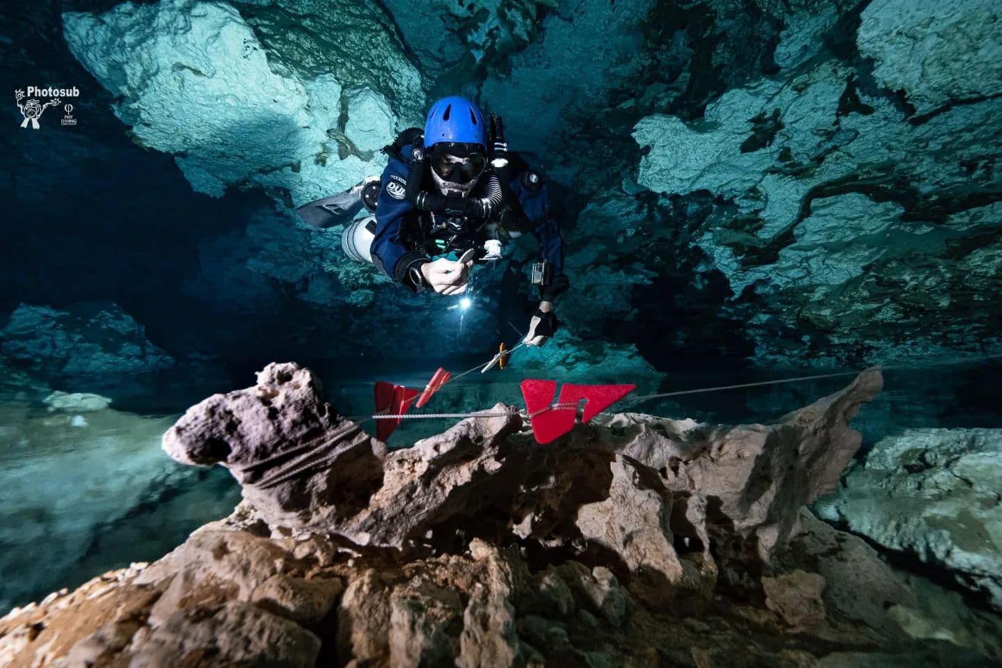 Cave diver with blue helmet placing red directional arrow markers on a guideline near a rock formation