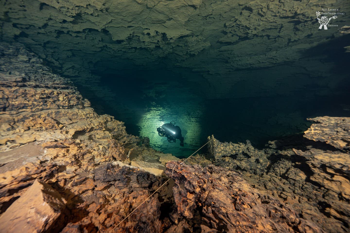 Lone diver following a guideline through a large dark cave chamber illuminated by a single primary light