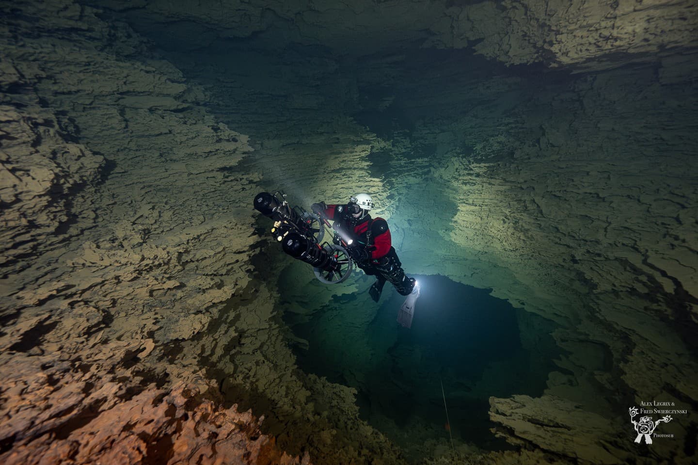 Cave diver in red drysuit with DPV underwater scooter and camera equipment navigating cave passage