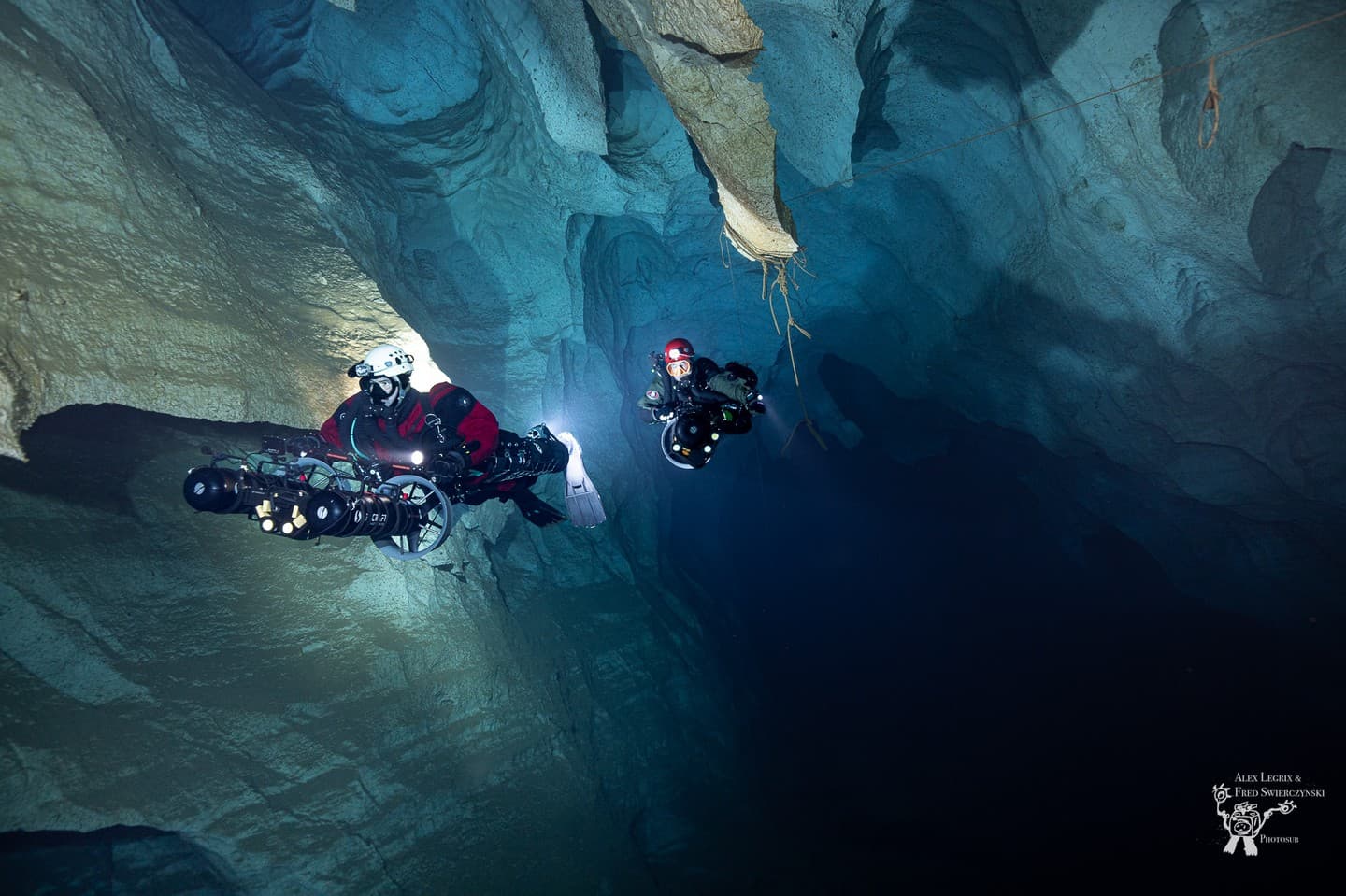 Two cave divers with underwater scooters in a large cave chamber with hanging stalactite formations