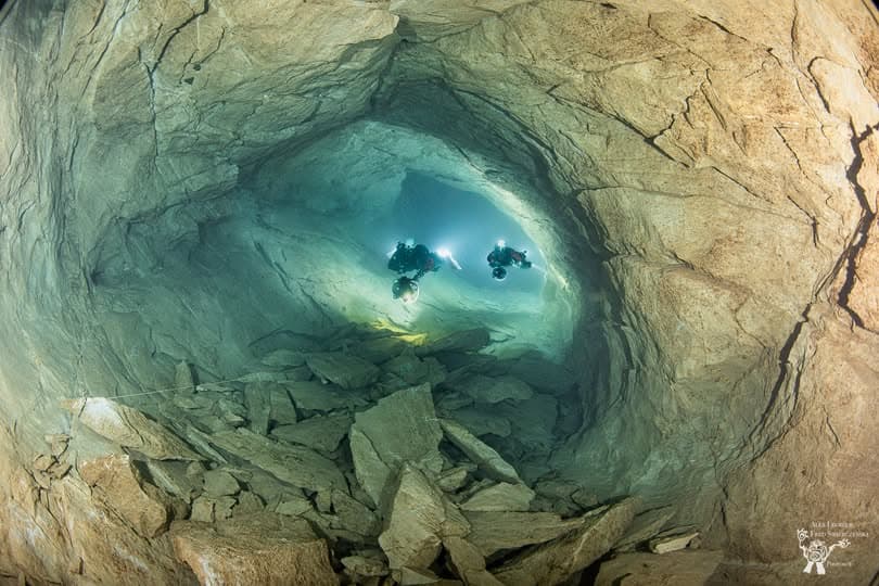Fish-eye lens view of two cave divers in a large rocky chamber with angular rock formations below