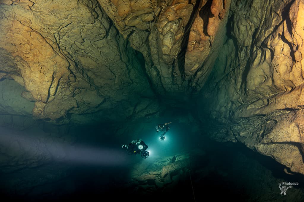 Two cave divers with DPVs illuminated by a dramatic shaft of light in a dark cave chamber