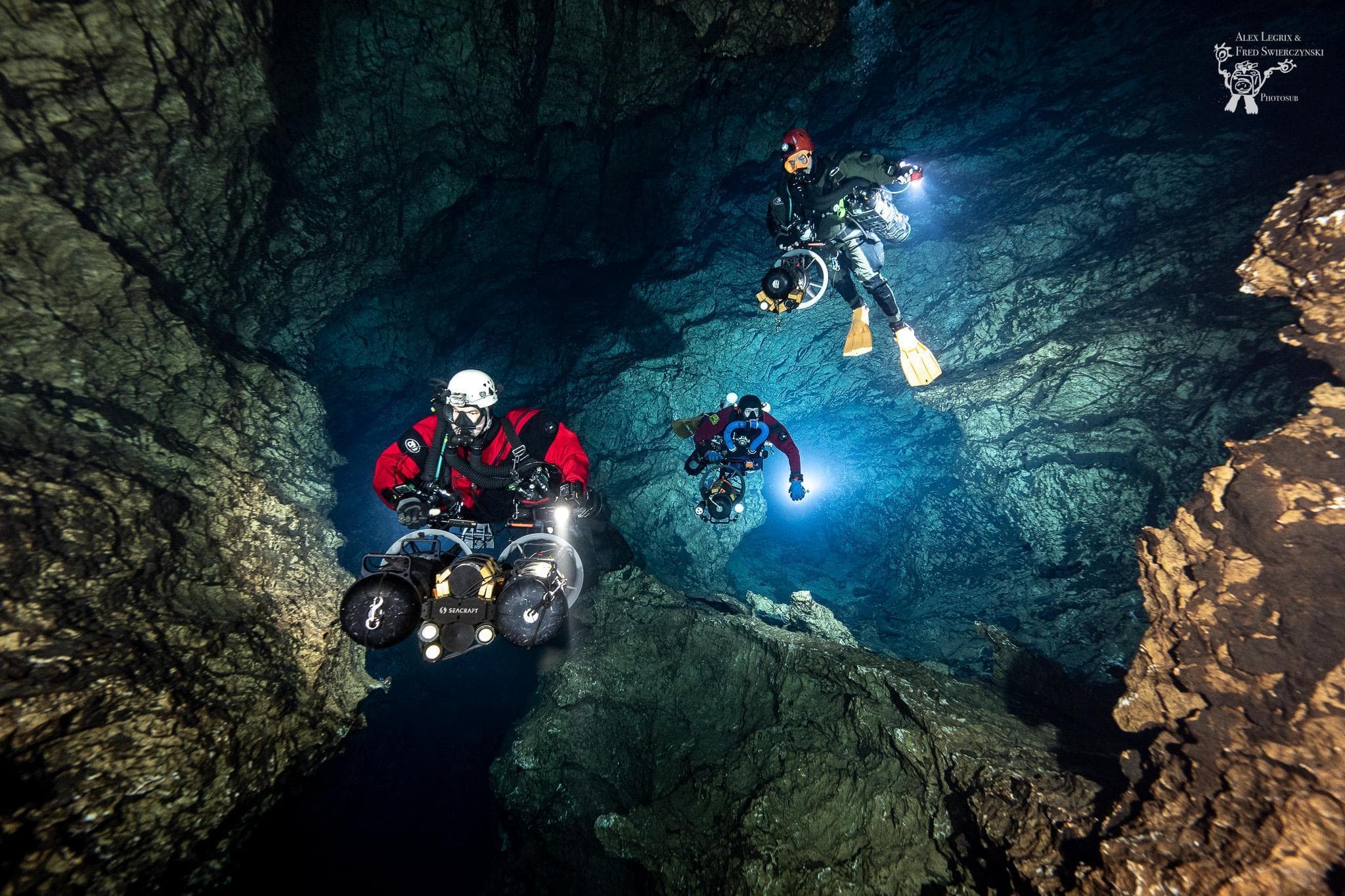 Three cave divers with underwater scooters navigating a large cave chamber with bright lights